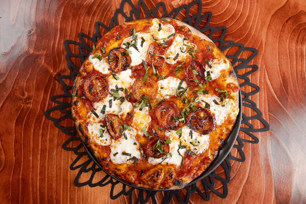 Pizza with mozzarella, roasted tomatoes, and basil on a black trivet on a wooden table.