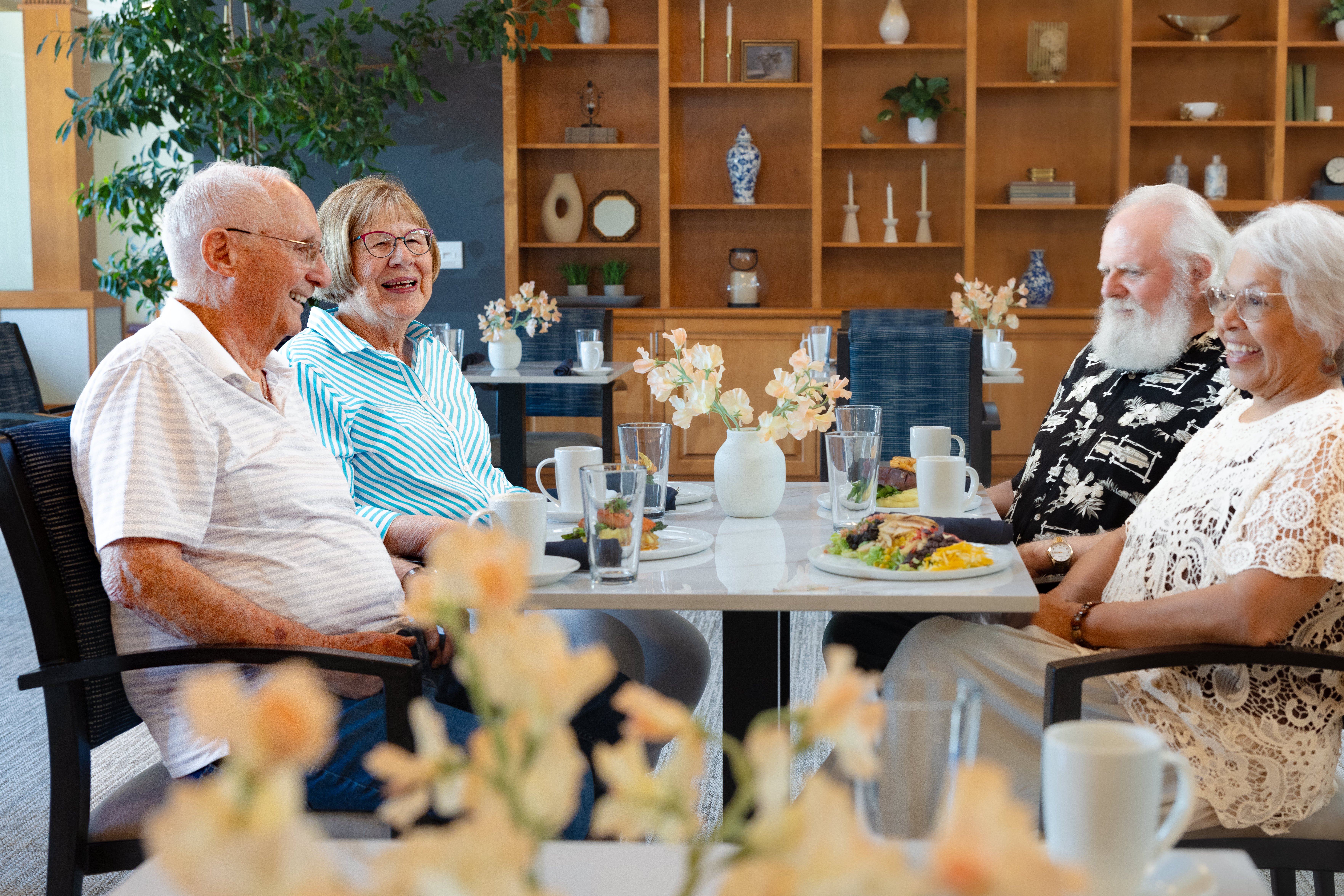 Five older adults smiling and sharing a meal together at a table in a bright community dining room.