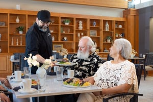 A chef serves food to an elderly couple at a dining table in the bistro