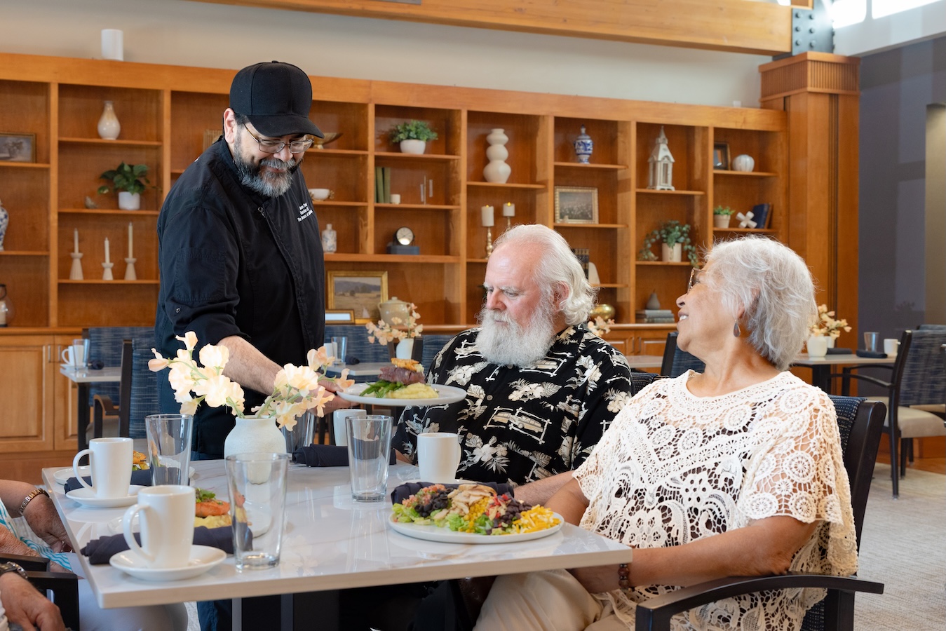 A chef serves food to an elderly couple at a dining table in the bistro