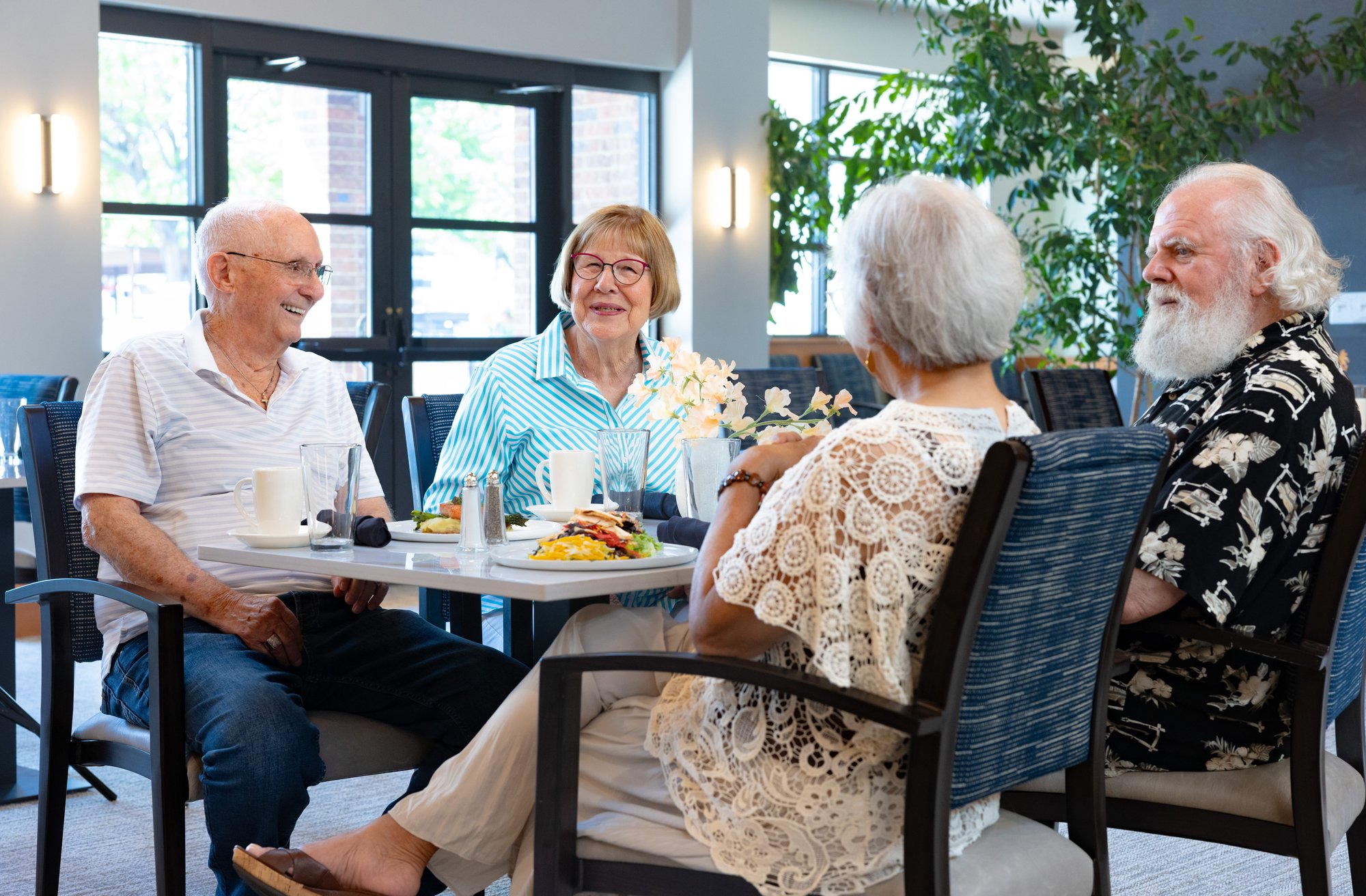 Four older adults enjoying a meal and conversation together at a dining table in a bright community dining room.