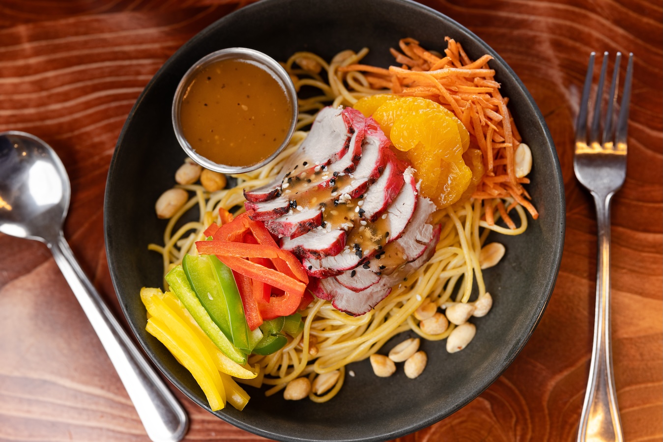 A bowl of noodles with sliced grilled meat, bell peppers, grated carrots, orange slices, peanuts, and a cup of sauce.