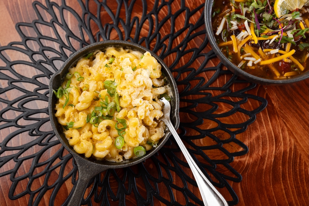 Macaroni and cheese and a bowl of chili on a decorative black trivet on a wooden table.