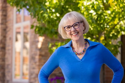Terri in a blue sweater posing outdoors, smiling with hands on hips. Behind her are trees and a building.