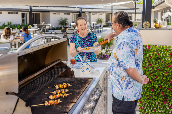 Senior man in a Hawaiian shirt placing a grilled kebab on a senior woman's plate at a barbecue