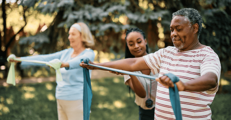 older adult man and woman using exercise bands outside with a trainer