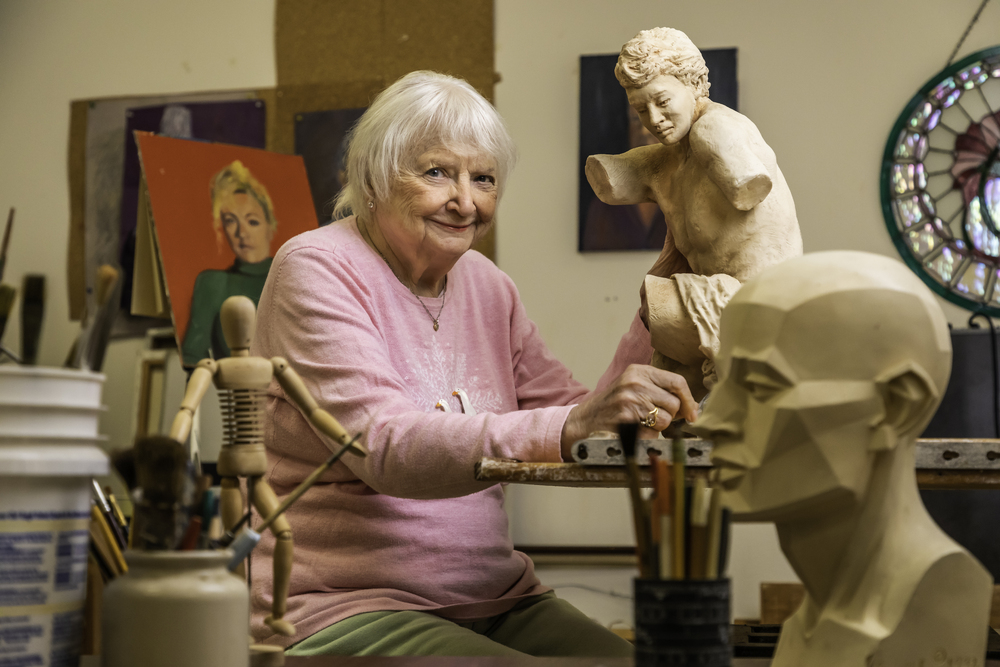 Woman artist in a pink sweater works on a wooden sculpture in her studio, surrounded by art tools, paintings, and sculptures.