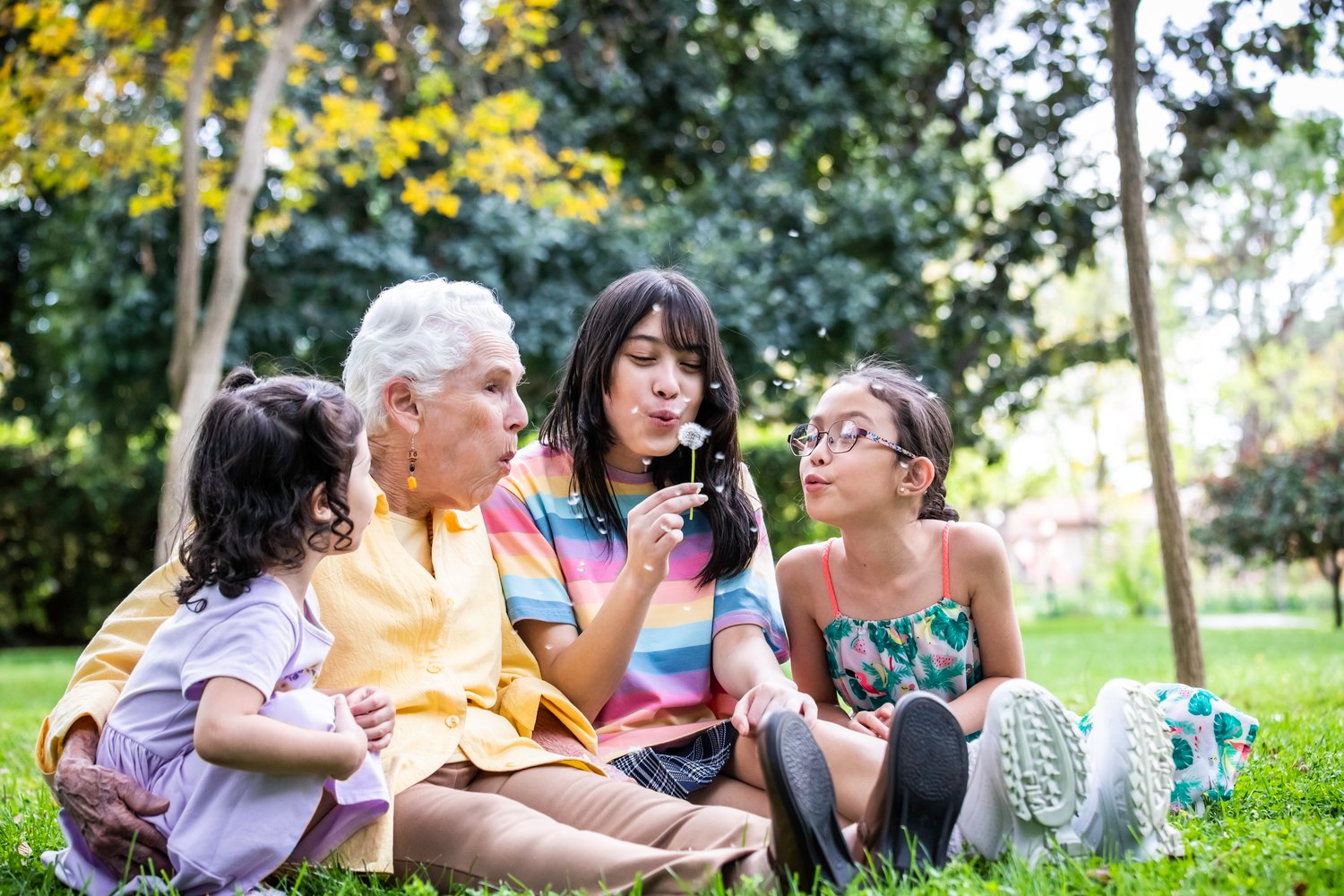 Grandmother sitting in park with grandchildren blowing on dandelion seeds.