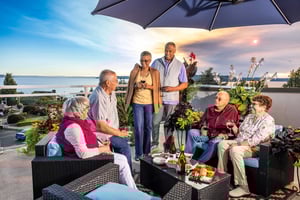 Residents gather on an outdoor terrace at Judson Park, enjoying conversation, drinks, and snacks with a waterfront view at sunset.