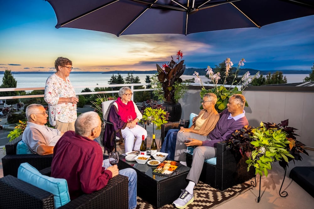 A group of friends enjoying a gathering on a balcony with a sunset view, surrounded by plants and snacks.