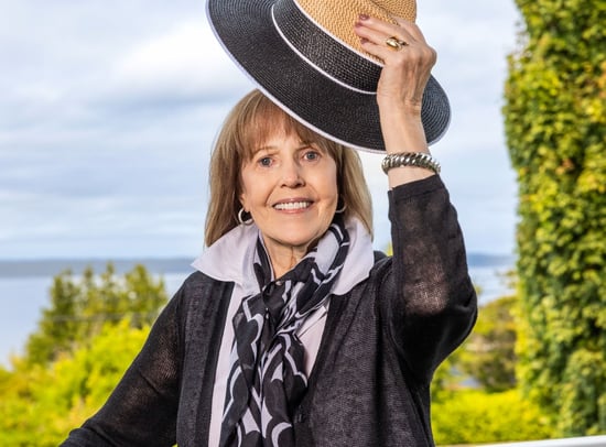 Linda smiling outdoors and holding a black and tan hat.