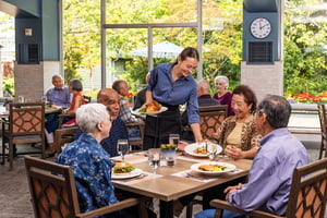 Residents enjoying a shared meal while a staff member serves food in a dining space at Judson Park
