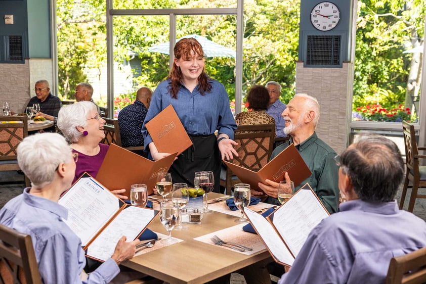 A server chats with residents seated around a dining table at Judson Park, as they review menus and enjoy a relaxed, social meal together.