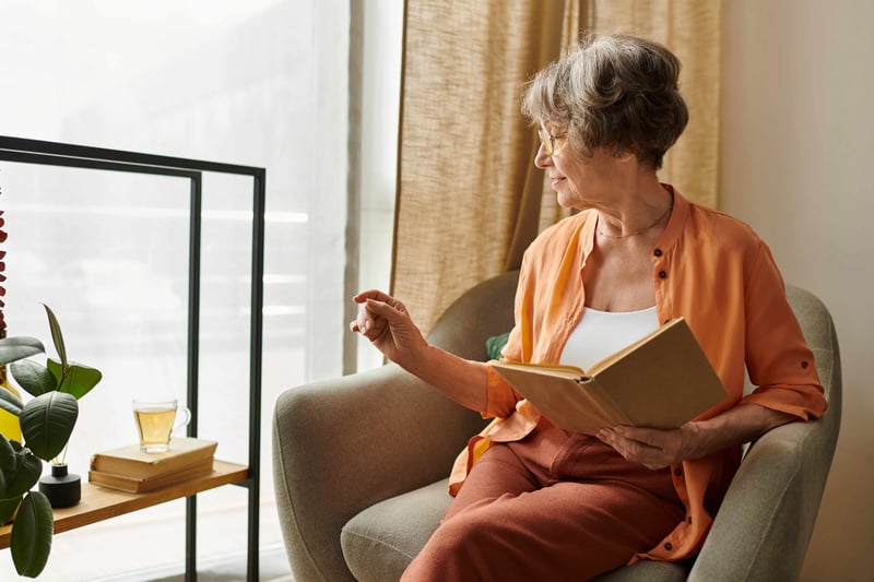 senior woman relaxing in a chair by the window with a book, reaching for a cup of tea on the shelf beside her