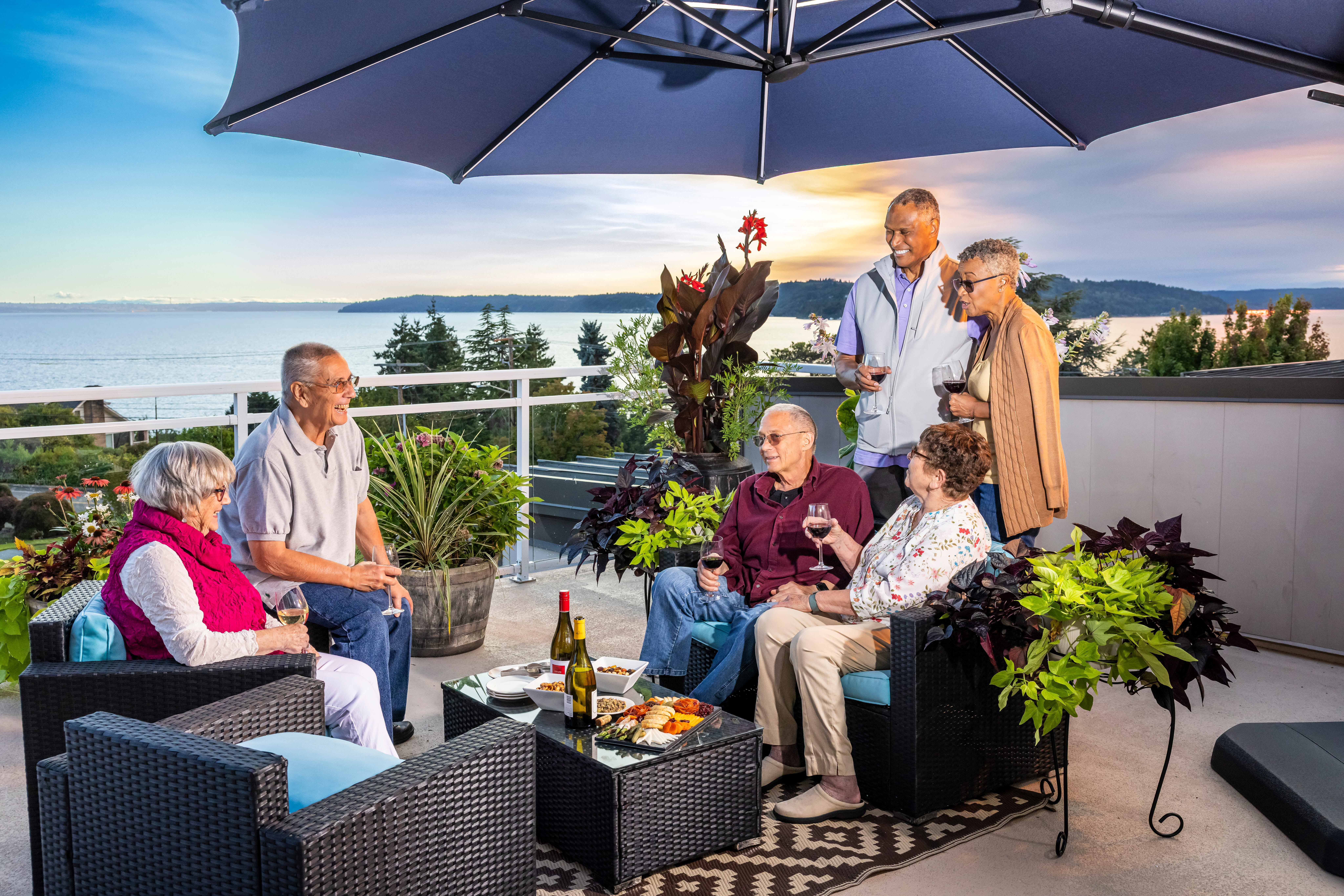 Group of six senior friends enjoying food and drinks on a rooftop balcony over the sunset