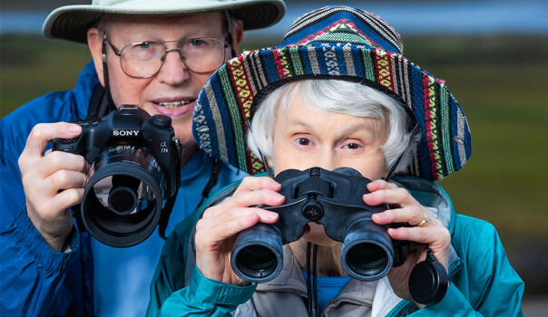 Woman with colorful hat uses binoculars, while man beside her holds a camera.