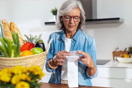 Senior woman reviewing a receipt in her kitchen with a basket of produce and bread on the counter