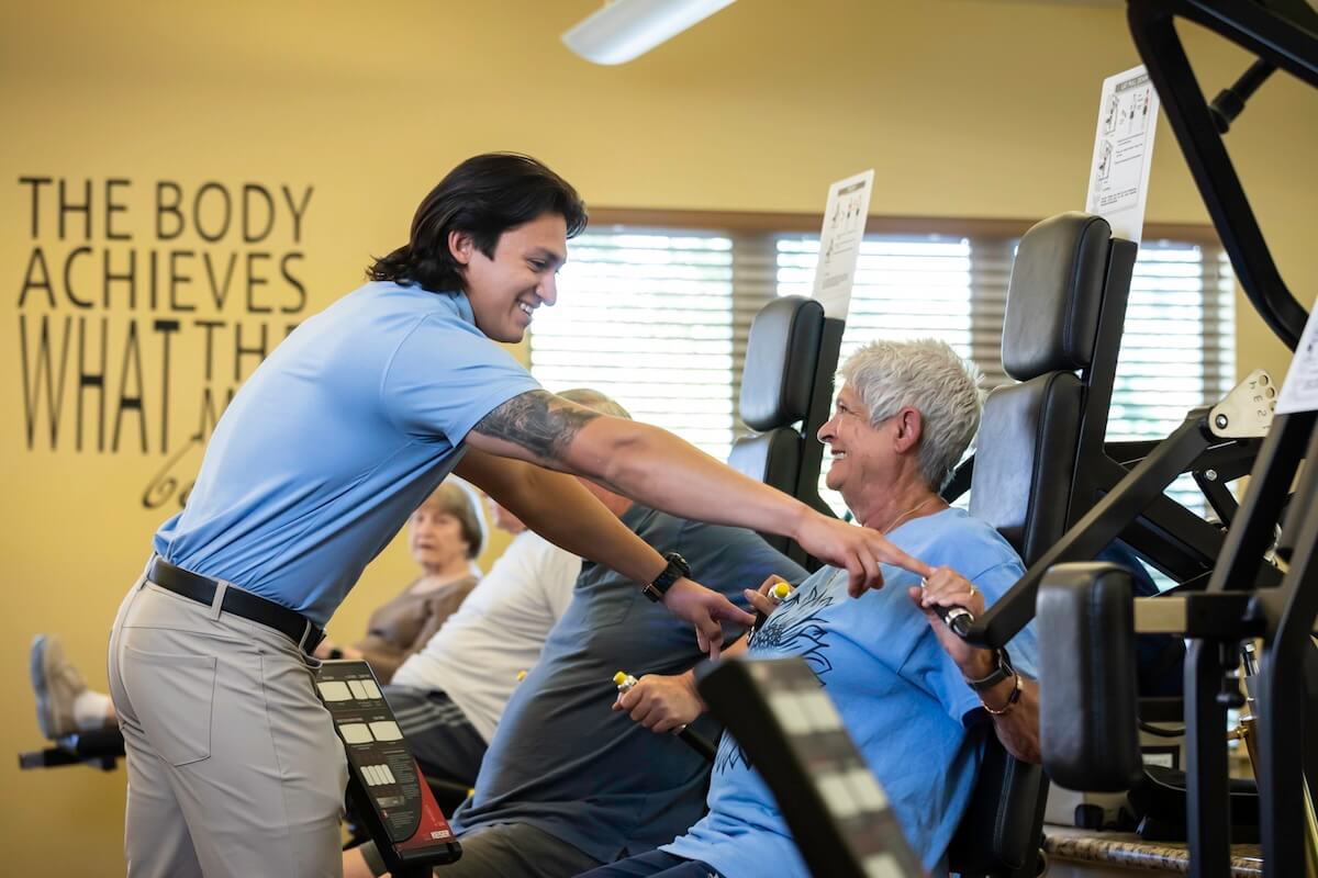 Senior woman using weight machine in fitness center with the assistance of personal trainer