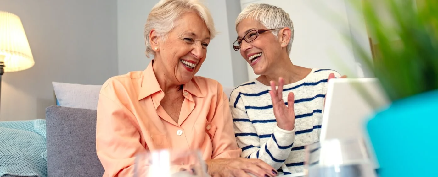 Two senior women sitting next to each other on couch while laughing
