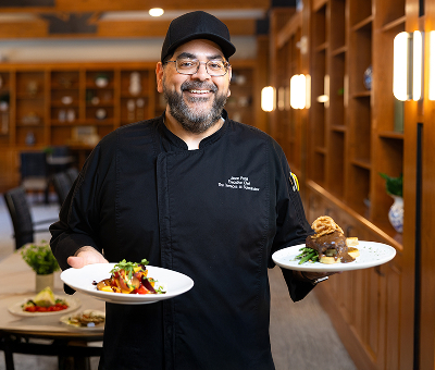 A smiling chef in a black uniform holds two plates of gourmet food in a warmly lit restaurant, conveying a sense of pride and culinary expertise.