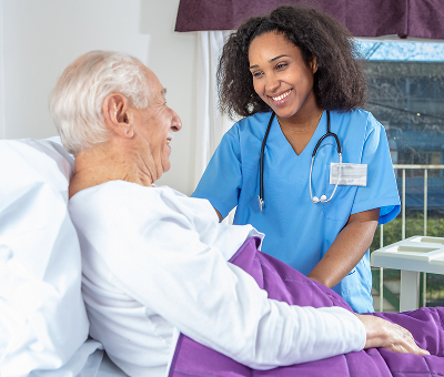 A smiling nurse in blue scrubs and a stethoscope attends to an elderly man in a hospital bed covered with a purple blanket
