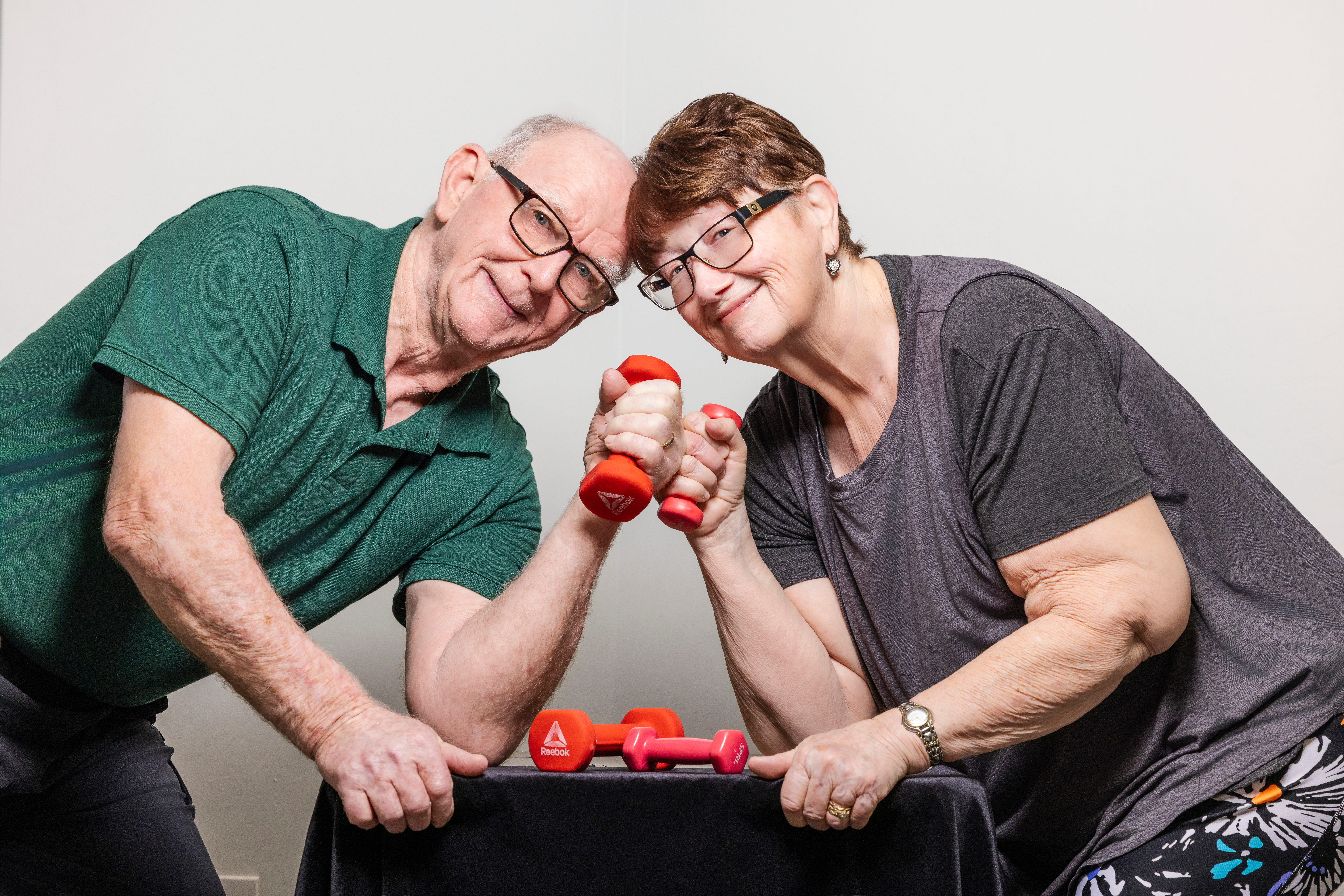 Ruth and Tom are lifting lightweight red dumbbells for a picture.