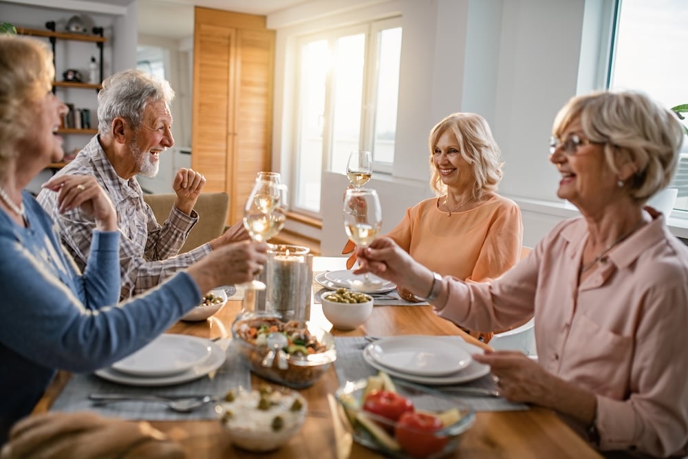 Four older adults smile and raise wine glasses around a dining table filled with food