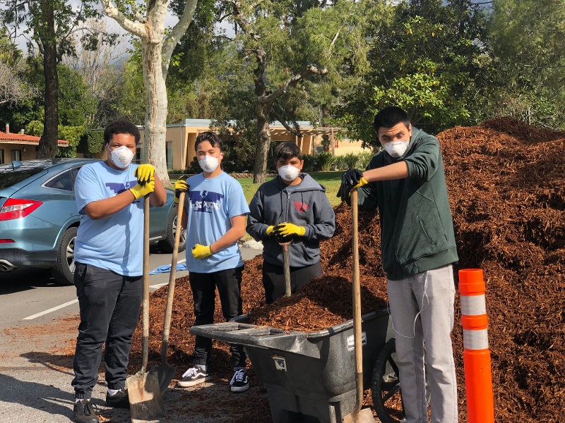 A group of four posing with tools, a wheelbarrow full of mulch, and a large pile of mulch behind them.