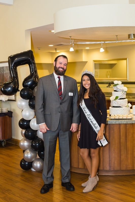 A man in a suit smiling next to a girl in a crown and sash that reads 