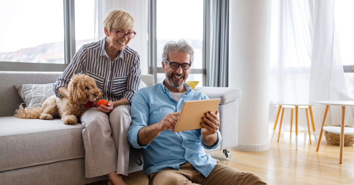 man and woman smiling on couch with their dog looking at a tablet