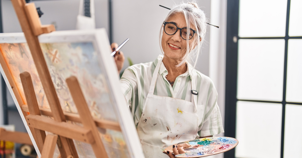 older woman in white apron painting on a canvas while holding a paint palette
