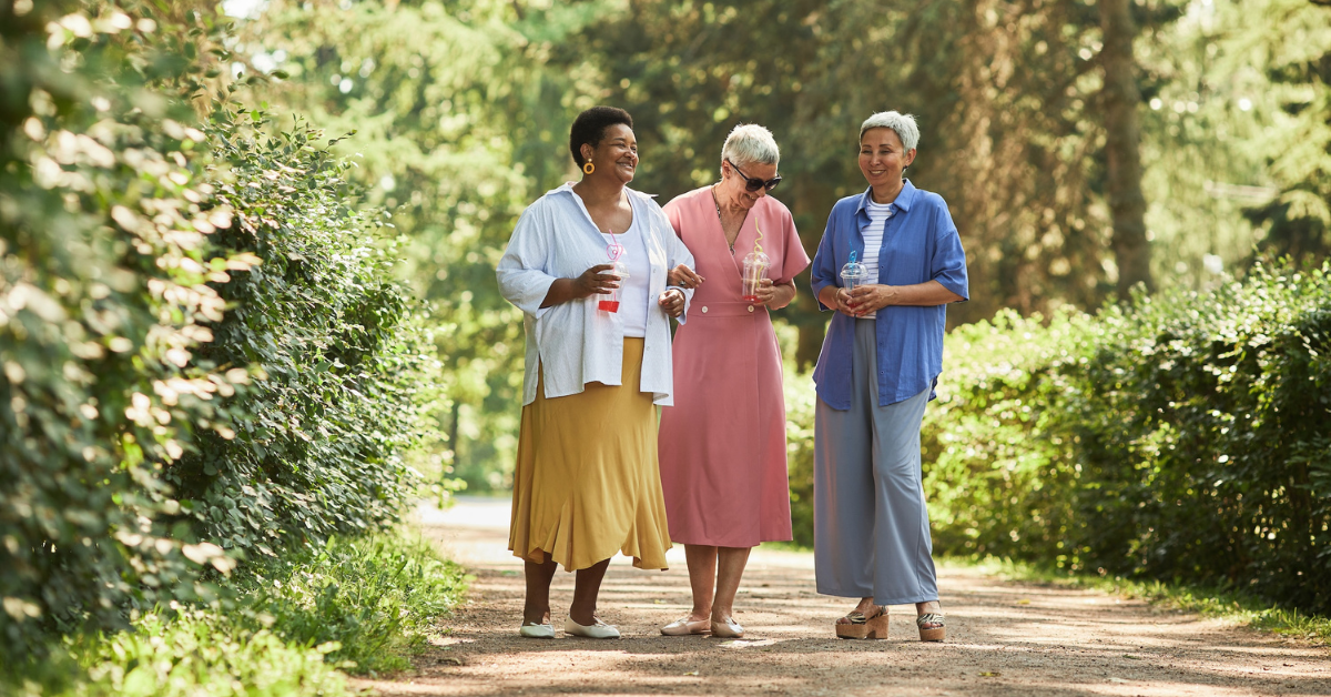 group of 3 senior women walking through a wooded path holding drinks and smiling