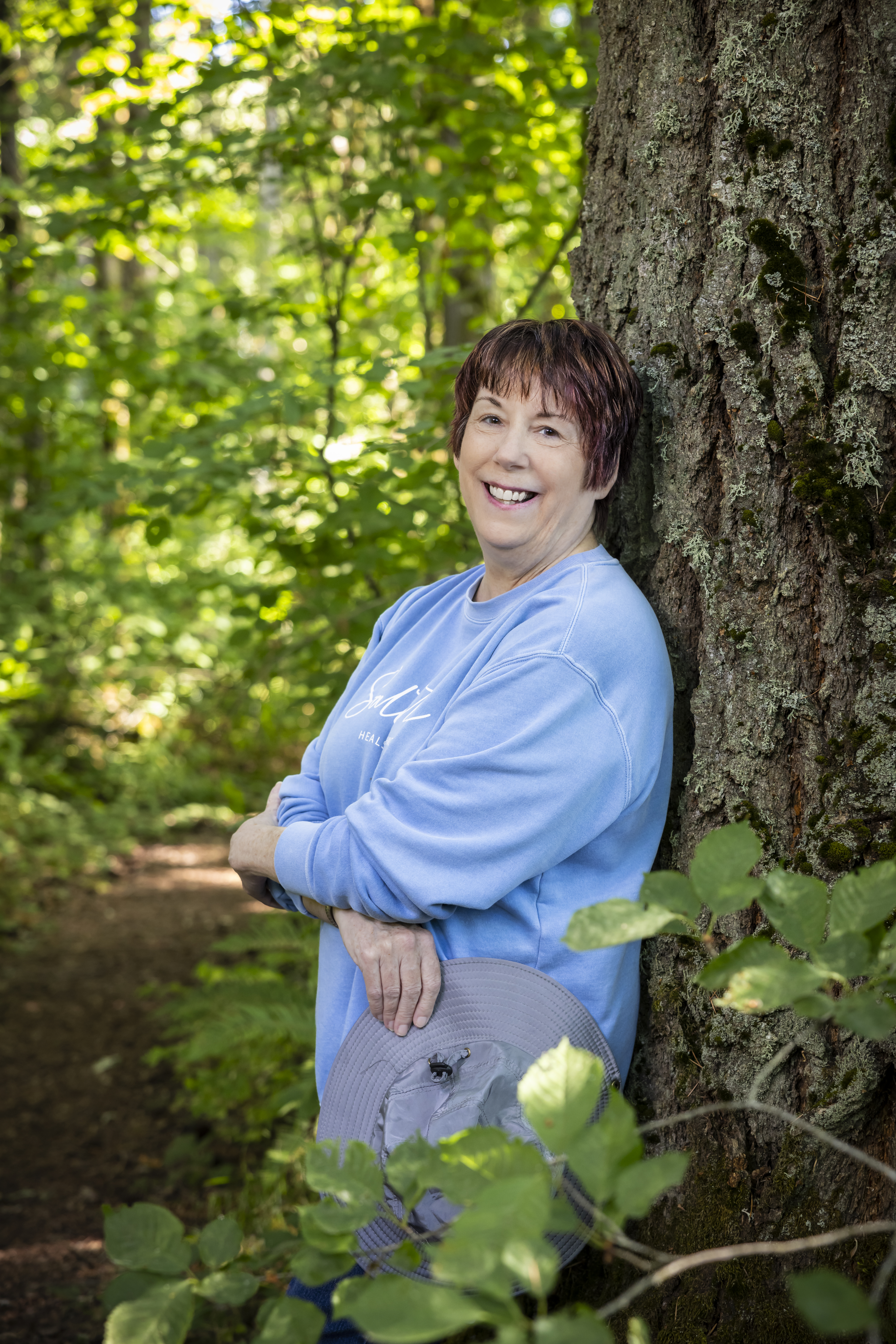Erin, a resident at the Hillside community, smiling and leaning against a tree wearing a blue shirt with arms crossed